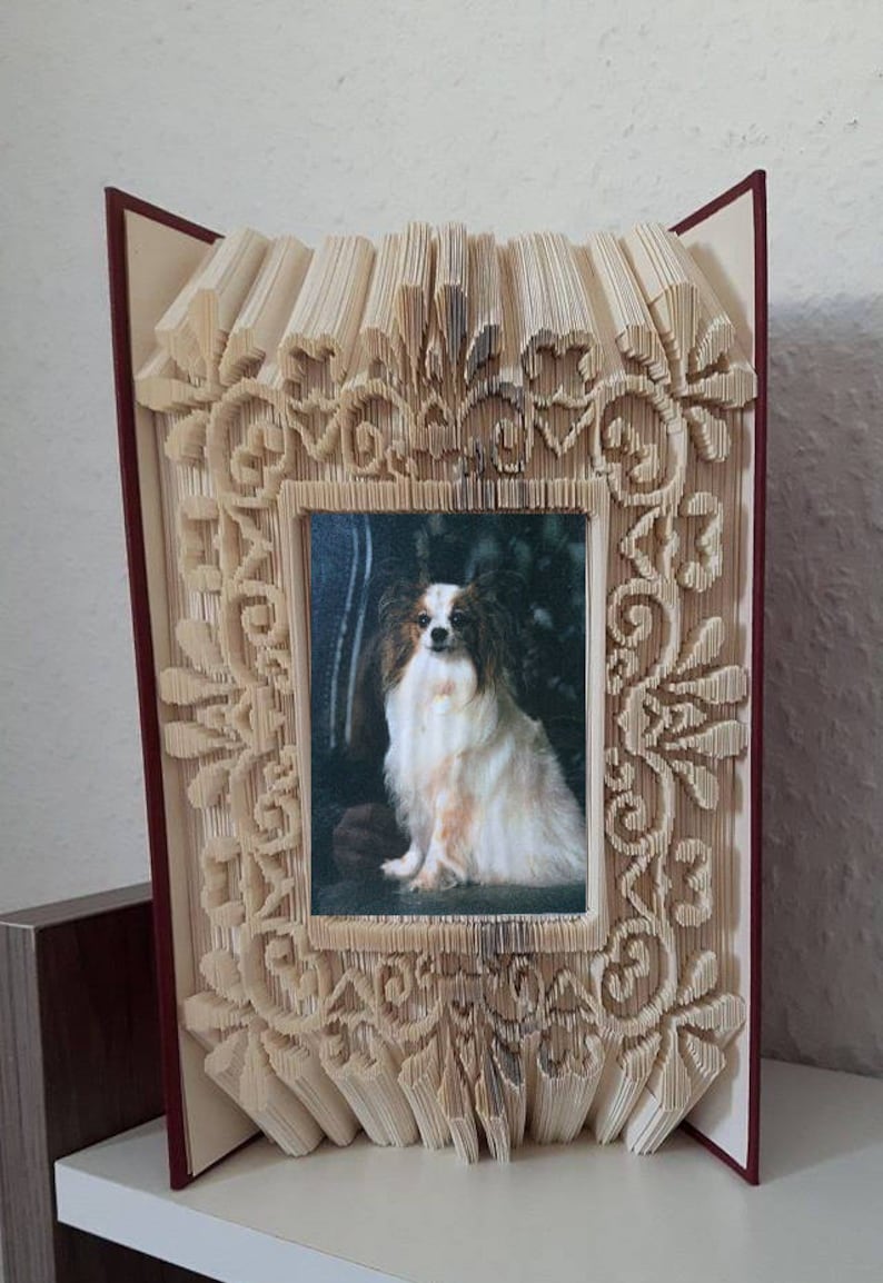 May include: A book art frame made from a book with pages folded into a decorative design. The frame is holding a photo of a papillon dog.