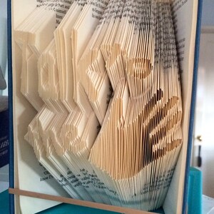 Op de afbeelding: Een boek met pagina's die zijn gevouwen tot een driedimensionaal ontwerp, waardoor een decoratief beeldhouwwerk ontstaat. Het boek is in blauw gebonden en heeft een bruin elastiek eromheen.