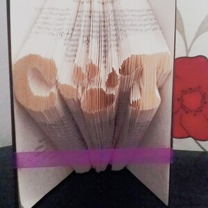 May include: A book with pages folded into the shape of the word "LOVE" in a white book with a purple ribbon. The book is standing upright on a black surface.