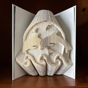 Puede incluir: Un libro blanco con las páginas dobladas en una escultura 3D de dos delfines saltando fuera del agua. La escultura se exhibe en un estante de madera.