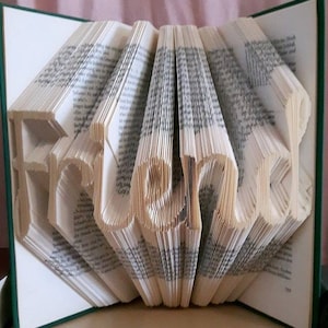Puede incluir: Una escultura de arte de libro con la palabra "Friend" doblada en las páginas. El libro está abierto y las páginas están dobladas para crear la palabra. El libro está sobre una superficie de madera.
