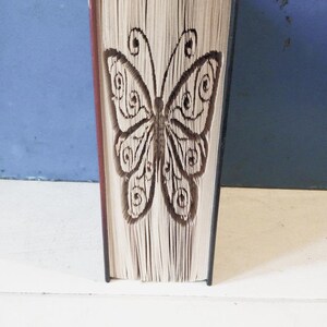 May include: A book with a butterfly design carved into the pages. The book is closed and standing upright on a white surface.