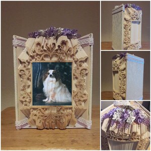 以下が含まれることがあります： 白い毛と茶色の毛の papillon の犬の写真が入った本の芸術フレーム。フレームは本の折り畳まれたページで作られており、紫色の花で飾られています。