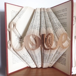 May include: A book with the word "LOVE" folded into the pages. The book is open and the pages are folded to create the word. The book is on a wooden surface.