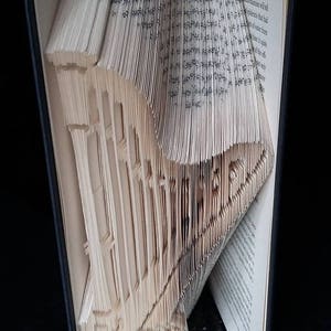May include: A book with pages folded into a 3D sculpture of a house. The book is open and the sculpture is visible from the side. The book is on a black surface.