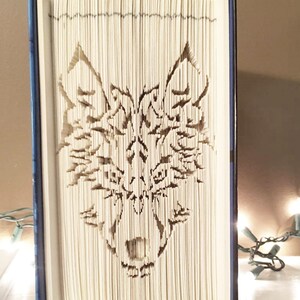 May include: A book with the pages folded to create a 3D sculpture of a wolf's head. The sculpture is made from the pages of a book, and the wolf's head is facing forward. The book is closed and standing upright on a shelf.
