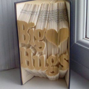 Può includere: Una scultura di arte di libro con le parole "Big Hugs" piegate nelle pagine. Il libro è in piedi su una superficie bianca.