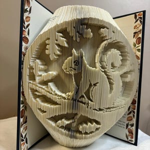 May include: A book art sculpture of a squirrel sitting on a branch, created by folding the pages of a book. The sculpture is made from a book with a brown cover and a floral design on the spine.