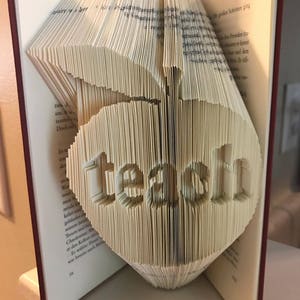 Peut inclure: Une sculpture d'art de livre du mot "teach" plié à partir des pages d'un livre. Le livre est ouvert et les pages sont pliées en une forme tridimensionnelle.