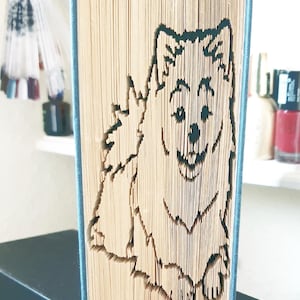 Puede incluir: Un libro con las páginas dobladas para crear una imagen 3D de un perro blanco. El perro está sentado con la cabeza inclinada hacia un lado y la boca ligeramente abierta. El libro está encuadernado con una cubierta azul.
