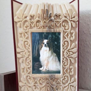 May include: A book art frame made from a book with pages folded into a decorative design. The frame is in the shape of a rectangle with a floral pattern. A photo of a white and brown papillon dog is inside the frame.
