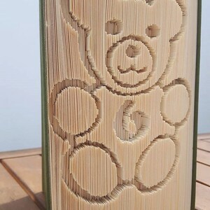 May include: A book with a green cover has been folded into the shape of a teddy bear. The pages are folded to create the bear's outline, and the bear's face, paws, and body are visible.