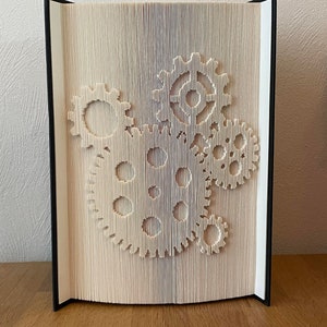 Könnte beinhalten: Ein weißes Buch mit einem 3D-Pop-up-Design aus Zahnrädern. Das Buch ist geschlossen und steht aufrecht auf einer Holzoberfläche.