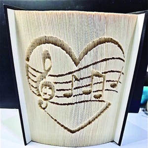 May include: A book art sculpture featuring a heart shape and musical notes carved into the pages. The book is bound in black fabric.