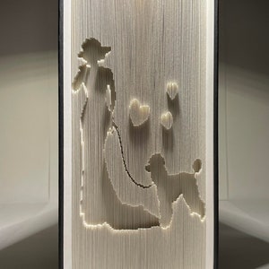 May include: A white book with a black cover has been folded into a 3D sculpture of a woman walking a poodle. The woman is wearing a hat and holding a leash. There are several hearts cut out of the pages.