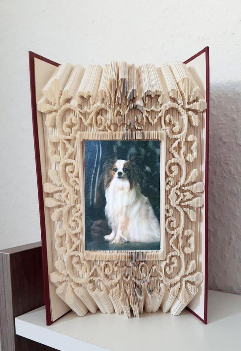May include: A book art frame made from a book with pages folded into a decorative design. The frame is in the shape of a rectangle with a picture of a white and brown dog inside.