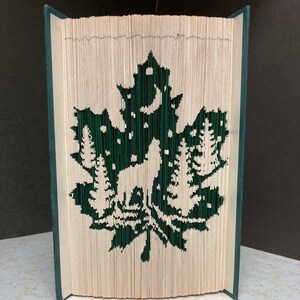 May include: A book art sculpture featuring a green silhouette of a maple leaf, a wolf, and trees. The book is folded and shaped to create a 3D effect.