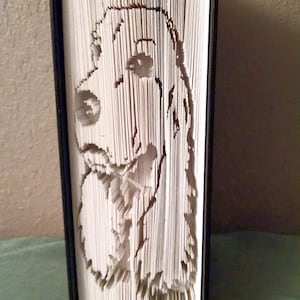 May include: A book with the pages folded to create a 3D image of a dog's head. The dog is white with brown markings and has floppy ears. The book is bound in black.
