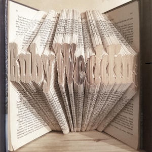 Könnte beinhalten: Ein Buch mit dem Wort "Happy Wedding" in die Seiten gefaltet. Das Buch ist geöffnet und die Seiten sind gefaltet, um das Wort zu bilden.