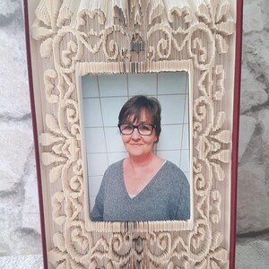 May include: A book with pages folded into a decorative frame, holding a photo of a woman with short brown hair wearing glasses. The frame is made of white paper and has a detailed floral design.