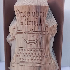 Può includere: Un libro piegato in una scultura 3D di una macchina da scrivere. Il libro è marrone con una copertina marrone. La scultura è intitolata "C'era una volta".