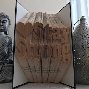 Può includere: Una scultura di arte libraria con le parole "Stay Strong" piegate nelle pagine. La scultura è realizzata con un libro con una copertina bianca e una rilegatura nera. La scultura è appoggiata su una superficie bianca.