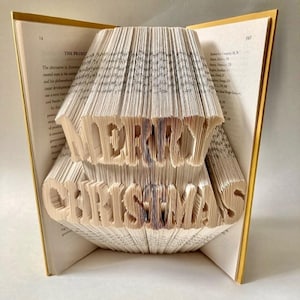 Könnte beinhalten: Ein Buch mit den Worten "Merry Christmas", die in die Seiten gefaltet sind. Das Buch ist geöffnet und die Seiten sind gefaltet, um einen 3D-Effekt zu erzeugen.