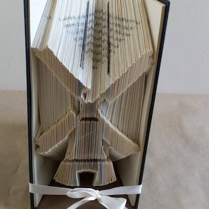 Puede incluir: Un libro con las páginas dobladas en forma de molino de viento. El libro está encuadernado en negro y tiene una cinta blanca atada alrededor.