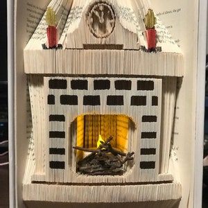 Op de afbeelding: Een boekvormige open haard met een klok erboven en een brandende kaars erin. De open haard is gemaakt van gevouwen pagina's uit een boek en heeft een zwart-wit baksteenpatroon.