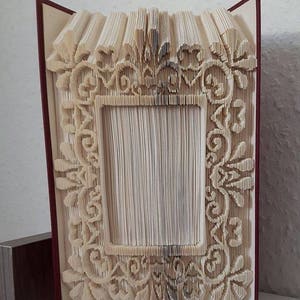 May include: A book art sculpture with a decorative frame design created by folding the pages of a book. The frame is made of intricate patterns and the book is bound in a burgundy cover.