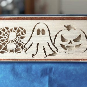 Op de afbeelding: Decoratief boek met het woord 'BOO' uitgesneden in de pagina's, met een spinnenweb, een spook en een pompoen. Het boek heeft een blauwe kaft en een bruine glitterrand, perfect voor Halloween-decoratie.