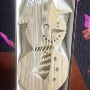 Peut inclure: Une sculpture d'art de livre représentant un bonhomme de neige réalisée en pliant les pages d'un livre. La sculpture est réalisée à partir d'un livre blanc avec une couverture marron.
