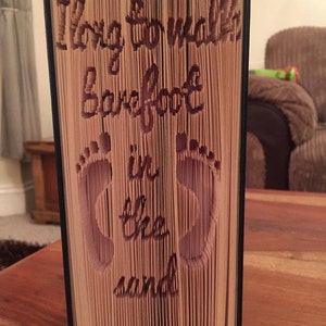 May include: A book with the words "I long to walk barefoot in the sand" carved into the pages. The book is folded in a way that creates a 3D image of two footprints.