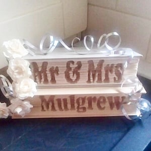 May include: A stack of books decorated with white flowers and silver ribbon. The top book is engraved with "Mr & Mrs Mulgrew".
