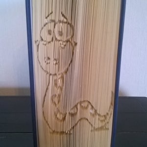 以下が含まれることがあります： 驚いた表情をした漫画のヘビの折り紙デザインが施された本。本は閉じられており、暗い表面に立てられています。
