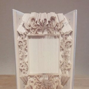 以下が含まれることがあります： ページが折り畳まれて装飾的なフレームの形になった白い本。フレームは複雑な紙のデザインでできています。