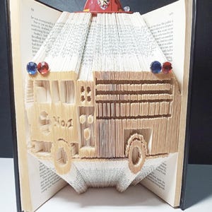 May include: A red fire engine crafted from the pages of a book. The engine is detailed with a red fire hat on top and blue and red gems on the sides.