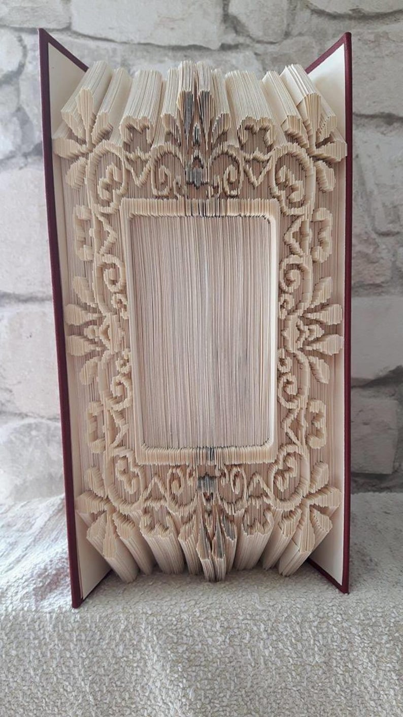 May include: A book with pages folded into a three-dimensional frame design. The frame is made of white paper and has a decorative, ornate pattern. The book is bound in red.