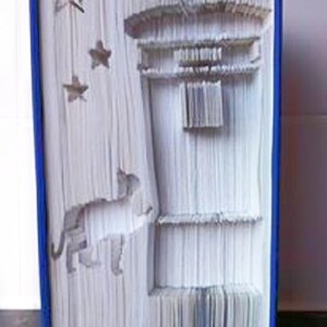 Könnte beinhalten: Ein Buch mit Seiten, die zu einem dreidimensionalen Design eines Katers, eines Vogels und eines Gebäudes mit Sternen im Hintergrund gefaltet sind. Das Buch ist in Blau gebunden.