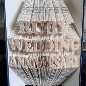 May include: A book with the words "First Wedding Anniversary" folded into the pages. The book is open and the pages are folded to create a 3D effect. The book is blue on the outside.