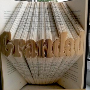 May include: A book with the word "Grandad" sculpted from its pages. The beige pages are folded to create the letters. A rubber band keeps the book open. The book is placed near a window.