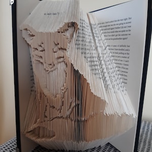 May include: A book art sculpture of a cat made by folding the pages of a book. The sculpture is made from a book with a black cover.