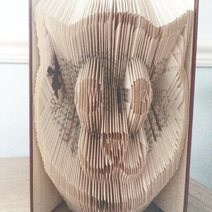 May include: A book with pages folded into the shape of an owl. The book is bound in brown leather and is standing on a wooden surface.