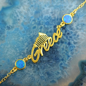 Puede incluir: Una pulsera dorada con la palabra "Greece" en cursiva y un pequeño diseño arquitectónico. La pulsera incluye tres piedras de ópalo azul. El diseño se destaca sobre un fondo azul.