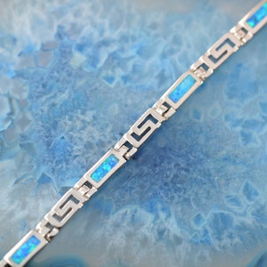 以下が含まれることがあります： ギリシャの鍵のデザインと長方形の青いオパールインレイが特徴のシルバーブレスレット。 ブレスレットは青い背景に表示され、シルバーとオパールのコントラストを際立たせています。 ブレスレットの長さは約18cmです。