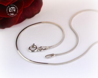 Puede incluir: Un collar de cadena de serpiente de plata con cierre de langosta. La cadena está colocada sobre una superficie blanca.
