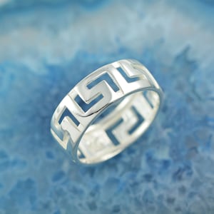 以下が含まれることがあります： ギリシャの鍵のデザインが施されたシルバーリング。リングは、薄くカットアウトされたパターンで金属のバンドで作られています。