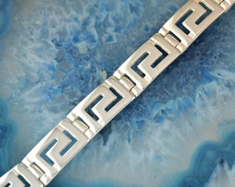 May include: A silver-toned bracelet with a Greek key design. The bracelet is composed of rectangular links, each featuring a repeating geometric pattern. The links are connected by smaller, rectangular pieces. The bracelet is displayed against a blue background.