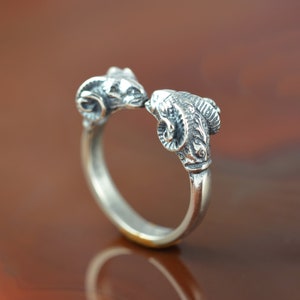 May include: A silver ring with two ram heads facing each other. The ram heads are detailed with horns and facial features.