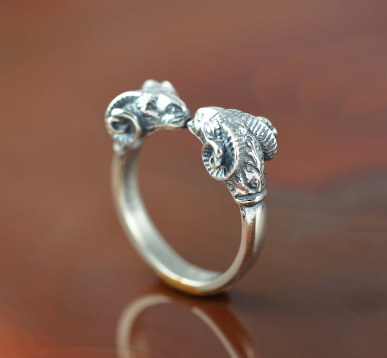 May include: A silver ring with two ram heads facing each other. The ram heads are detailed with horns and facial features.